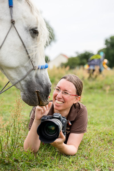 Fotografin_Jessica-Freymark2_Foto(c)AndreasBender Foto von Jessica Freymark wie sie im Gras liegt mit ihrer Kamera und lachend Kontakt zu einem neugierigen Pferd aufnimmt.