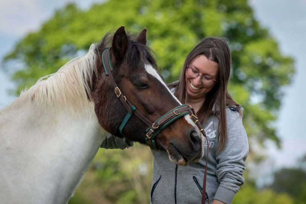 Autorin Jessica Freymark und ihr Pflegepferd Janek in einem innigen Moment. Janek trägt noch den Kappzaum, schließt die Augen und spitzt genüsslich die Oberlippe, während Jessica seinen Hals krault.